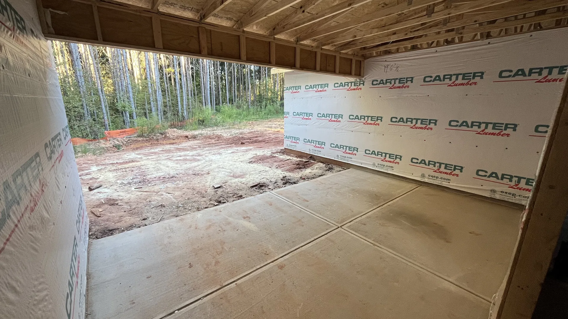 View beneath exposed overhead framing at the Contemporary Ranch, showing housewrap, the concrete slab, and the wooded site — York County, SC